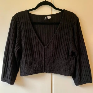 Svart ribbad topp - Säljer en snygg svart ribbad topp från H&M. Den har en djup v-ringning och knäppning framtill. Toppen är i mycket bra skick och har trekvartsärmar.