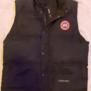 Säljer en svart dunväst från Canada Goose i mycket bra skick. Västen har en klassisk design med knappar och dragkedja framtill samt en ikonisk logga på bröstet. Perfekt för kyliga höst- och vinterdagar.