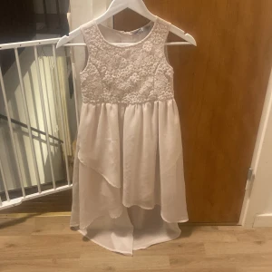 Vit ärmlös klänning med spetsdetaljer - Säljer en superfin vit klänning med spetsdetaljer på överdelen. Den är ärmlös och har en elegant asymmetrisk kjol som är längre bak. Perfekt för sommarens fester eller studentfiranden! Klänningen har en dragkedja i ryggen för enkel påklädning.