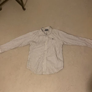 Randig skjorta från Ralph Lauren - Säljer en stilren och klassisk randig skjorta från Ralph Lauren. Skjortan är vit med tunna svarta ränder och har långa ärmar. Perfekt för både vardag och fest! Den har en broderad logga på bröstet och knappar framtill. Passar bra till både jeans och chinos.