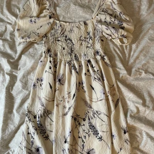 Blommig klänning med puffärmar - Superfin blommig klänning i ljus färg med puffiga kortärmar och smockdetaljer på överdelen. Perfekt för sommarens fester eller en mysig vårdag. Den har ett romantiskt mönster med blommor i mörka toner och en luftig passform.