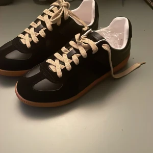 Svarta sneakers med beige snörning - Snygga svarta sneakers i mycket bra skick med beige snörning och brun sula. Perfekta för vardagsbruk eller en avslappnad dag ute. De har en stilren design med en kombination av mocka och syntetmaterial. Passar bra till både jeans och shorts!