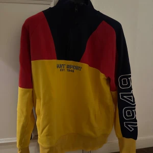 Flerfärgad tröja från Gant - flerfärgad tröja från Gant med texten 'GANT SPORT EST. 1949' på framsidan. Tröjan har en kombination av gult rött och mörkblått. Passar till höst samt vår. Tröjan är av storleken XL