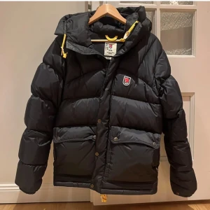 Svart dunjacka från Fjällräven - Säljer en svart dunjacka från Fjällräven i  XS. Jackan har en pufferstil med långa ärmar och en praktisk huva. Den är perfekt för kalla vinterdagar och har både dragkedja och knappar för extra skydd mot kylan. Fickor framtill för förvaring. 🖤fråga bara om du skulle vilja ha fler bilder🩷jacka är som ny