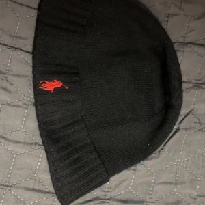 Svart mössa med röd broderad logga - Snygg svart mössa med en röd broderad logga på sidan. Den är ribbad och ser riktigt varm och skön ut för kyliga dagar. Perfekt för både höst och vinter. Passar till de flesta outfits och håller dig stilren och varm.