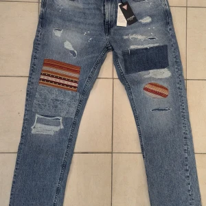 Blå jeans med patchwork från Guess - Säljer ett par snygga blå jeans från Guess med coola patchwork-detaljer och slitningar. De har en straight fit och är perfekta för en avslappnad stil. Jeansen har en klassisk femficksdesign och är i nytt skick. Perfekta för både vardag och fest!Nya aldrig använda. Taggar kvar. Köpta från Zalando. 