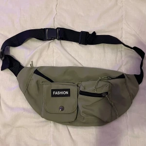 Grön magväska med flera fack - Säljer en grön magväska med justerbart band och flera praktiska fack. Väskan har en stor dragkedja upptill och mindre fack framtill, perfekt för att hålla ordning på dina saker. Märkt med 'FASHION' på framsidan. Perfekt för vardagsbruk eller resor! Oanvänd säljs för 40