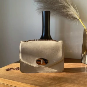 Beige axelväska med kedja - Säljer en stilren beige axelväska i syntetmaterial. Väskan har en elegant kedja (lite råstig) som axelrem och ett snyggt vridlås i guld på framsidan. Perfekt för både vardag och fest! 👜✨