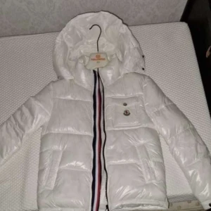 Vit dunjacka från Moncler - Säljer en snygg vit dunjacka från Moncler i mycket bra skick. Jackan har en quiltad design med en cool randig dragkedja i rött, vitt och blått. Den har en praktisk huva och en knappdetalj på bröstfickan. Perfekt för vintern och håller dig varm och stilren! ❄️