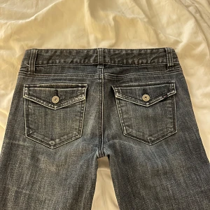 Mörkblå jeans med fickdetaljer - Säljer ett par mörkblå jeans i bra skick. De har en bootcut-stil och är lågmidjade. Baktill finns snygga fickor med knappar som ger en extra touch. Perfekta för både vardag och fest! Midja: 36cm o innerben: 80cm 