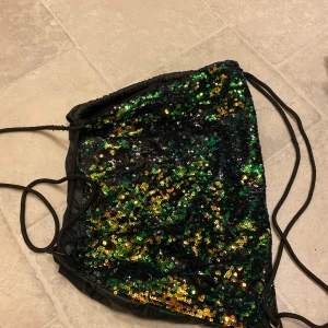 Svart paljettprydd topp - Säljer en glittrig svart tugväska med gröna och guldiga paljetter. Perfekt för festliga tillfällen när du vill sticka ut och glittra lite extra! 🖤✨ den funkar också att ha som glittrig gympapåse.