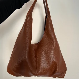 Väska - Säljer denna fina bruna väska i läder-liknande material från shein. Den är rymlig och i bra skick då jag endast använt den ett fåtal gånger. Man får även till en liten matchande necessär. Säljer den för 150kr.