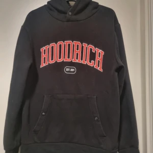 Svart hoodie från Hoodrich - Säljer en svart hoodie från Hoodrich med röd och vit text på framsidan. Den har en stor ficka fram och en luva med tryck. Perfekt för en casual look eller streetwear-stil. Använd men i bra skick !,lite avskaft på ena vita märket bild finns 