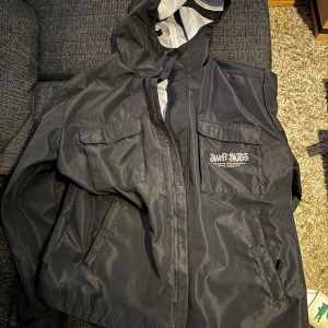 Svart vindjacka från Sweet Skateboard - Säljer en svart vindjacka från Sweet Skateboard i bra skick. Den har en praktisk huva och dragkedja framtill. Perfekt för blåsiga dagar och har en cool stil med logga på bröstet. Passar både till vardags och lättare utomhusaktiviteter. Size s passar m