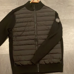 Svart Canada Goose cardigan (black label) - Säljer en snygg svart jacka från Canada Goose i mycket bra skick. Den har en quiltad framsida med dragkedja och ett stilrent märke på ärmen. med långärmade ärmar och en normal passform. En riktigt schysst jacka för alla tillfällen!