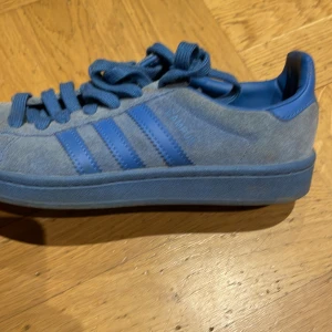 Blå Adidas Campus sneakers - Säljer ett par blå Adidas Campus sneakers i bra skick. Skorna har en snygg blå mockaöverdel med de klassiska tre ränderna i en mörkare blå nyans. De har snörning och en bekväm sula som passar perfekt för vardagsbruk. Perfekta för både vår och höst!