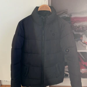 Svart pufferjacka från NLY Trend - Säljer en svart pufferjacka från NLY Trend i bra skick. Jackan har en dragkedja framtill och långa ärmar med ribbade muddar. Perfekt för kyliga höst- och vinterdagar. Den är både stilren och varm, vilket gör den till ett måste i garderoben! OBS den där lilla ”hängaren” på jackan har gått av. (Se bild)