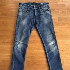 Blå Replay Anbass jeans - Säljer ett par snygga blå Replay Anbass jeans i bra skick. De har en cool slitning på ena benet och klassisk femficksdesign. Perfekta för en casual look och passar till det mesta. Midjan är normal och de har en bekväm passform.