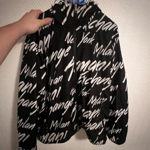 Svart och vit vindjacka från Armani Exchange - Säljer en cool svart vindjacka med vita textmönster. Jackan har en dragkedja framtill och är perfekt för höst och vår. Den är långärmad och har en avslappnad passform. Perfekt för dig som vill sticka ut lite i mängden! Sällan använd och i jättefint skick. Ställ gärna frågor angående bilder osv