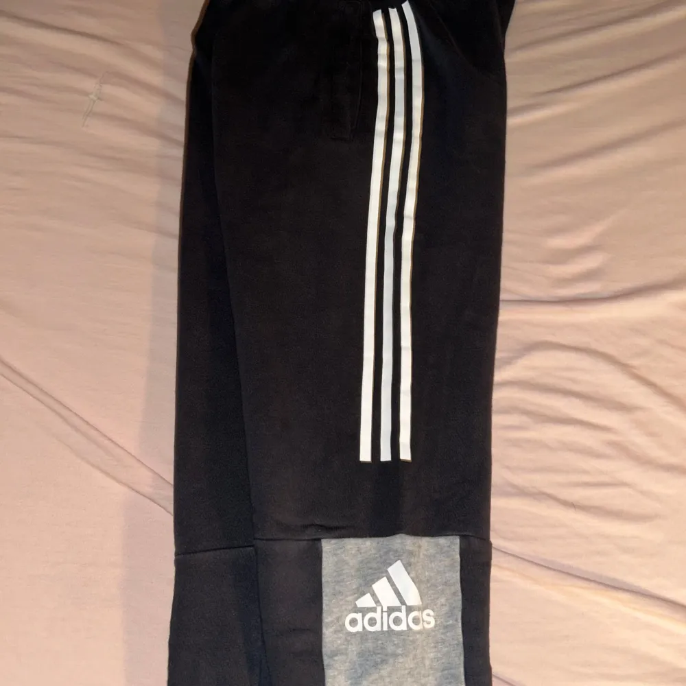 Säljer ett par snygga mjukisbyxor från Adidas i svart och grått. De har de klassiska tre vita ränderna längs sidorna och en grå sektion med Adidas-loggan på benet. Perfekta för träning eller chill hemma. Superbekväma och i bra skick!. Jeans & Byxor.