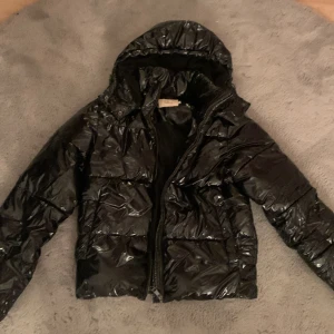 Svart pufferjacka från Nelly - Säljer en snygg svart pufferjacka från Nelly i storlek S. Jackan har en glansig finish och dragkedja framtill. Perfekt för kalla dagar med sin varma och vadderade design. Den har en huva för extra skydd mot väder och vind. Passar perfekt till både vardag och fest!
