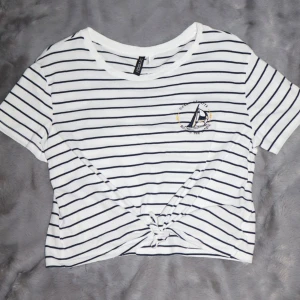 Randig t-shirt med båtbroderi - Vit och marinblå randig t-shirt. Den har ett broderi av en segelbåt på bröstet och en knutdetalj framtill. Använd ett fåtal gånger.