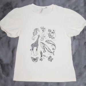 Vit t-shirt med djurmotiv - Vit t-shirt med djurmotiv i svart. T-shirten är kortärmad och gjord i mjukt bomullsmaterial. Den har puffärmar. T-shirten är i bra skick!