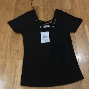 Svart topp med snörning från Inside - Snygg svart topp från Inside i storlek M. Den har en snörning framtill. Perfekt för både vardag och fest. Toppen är i nytt skick med etikett kvar.