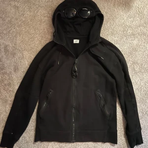 C.P. Company hoodie  - Säljer en cool svart hoodie från C.P. Company med inbyggda glasögon i huvan. Tröjan är köpt ifrån NK och säljs nu på grund av att den inte kommer till användning längre.