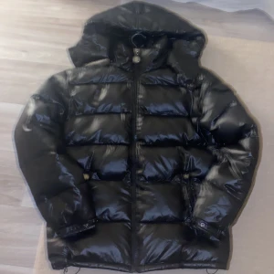 Svart pufferjacka med huva - Säljer en svart pufferjacka i mycket bra skick. Den är perfekt för vintern med sin quiltade design och långa ärmar. Jackan har en praktisk dragkedja och en justerbar huva. Den är gjord av glansigt polyester-material och har en cool ficka med knappdetaljer. Perfekt för att hålla sig varm och stilren i kylan!