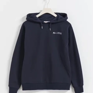 Mörkblå hoodie från Gina Tricot - Säljer en snygg mörkblå hoodie från Gina Tricot med texten 'MA CHÉRIE' tryckt på både fram- och baksidan. Den har en mysig huva och långa ärmar, perfekt för kyliga dagar. Materialet är mjukt och bekvämt, idealiskt för vardagsbruk. Passar perfekt för höst och vinter! De små metall sakerna på snörena har ramlat av en gång men limmat dem så de är inte helt säkert att de sitter jättebra.