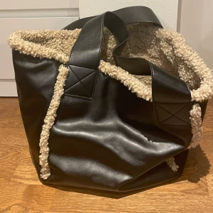 Svart axelväska med teddyfoder - Superfin svart väska i skinn med mysigt beige teddyfoder. Perfekt för höst och vinter! Väskan har en praktisk dragkedja och rejäla handtag. Den är rymlig (får plats med laptopp och annat) och passar perfekt till vardagen. Väskan är oanvänd. Nypris: 499
