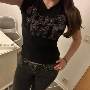 Svart t-shirt med tryck - Säljer en svart t-shirt med ett coolt tryck i lila och rosa nyanser. T-shirten har korta ärmar och är i bra skick. Perfekt för en avslappnad stil eller en utekväll. Passar bra med jeans eller kjol!