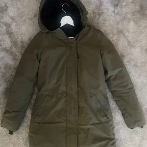 Olivgrön parkajacka från Canada Goose - Säljer en olivgrön parkajacka från Canada Goose i mycket bra skick. Jackan är perfekt för kalla vinterdagar med sin varma och tåliga design. Den har en långärmad stil med dragkedja och en praktisk huva. Märkets logga syns tydligt på ärmen. Perfekt för dig som vill hålla värmen med stil!