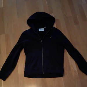 Svart hoodie Lyle & Scott - Snygg svart hoodie från Lyle & Scott med dragkedja framtill. Perfekt för en avslappnad stil. Den har en klassisk design med huva och långa ärmar. Tveka inte på att skriva om ni har några och funderingar