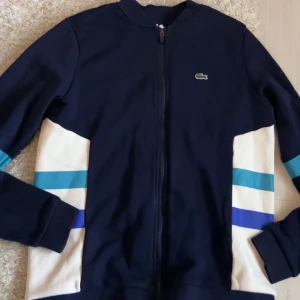 Blå och vit zip tröja från Lacoste för barn. - Säljer en trendig blå och vit zip tröja från Lacoste med dragkedja framtill. Tröjan har långa ärmar och  färgblock i blått och turkost på sidorna. Perfekt för en stilig look eller en avslappnad dag ute. Passar bra till alla årstider.
