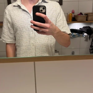 Vit och blå mönstrad skjorta från J.Lindeberg - Säljer en stilren vit skjorta med blått mönster från J.Lindeberg. Skjortan har korta ärmar och knappar framtill. Perfekt för en avslappnad look. Använt 1 gång 