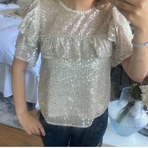 Glittrig topp med volanger - Säljer en superfin glittrig topp i beige med volanger. Den har korta ärmar och är perfekt för festliga tillfällen. Den är knappast använd och har inga defekter!❣️