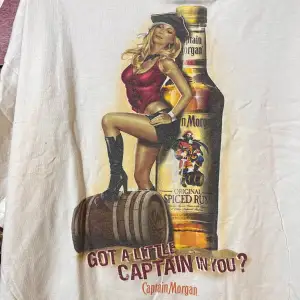 Säljer en vit t-shirt med ett coolt Captain Morgan-tryck. Den har en bild av en flaska och en kvinna i röd topp och svarta stövlar. Perfekt för en avslappnad dag eller fest.