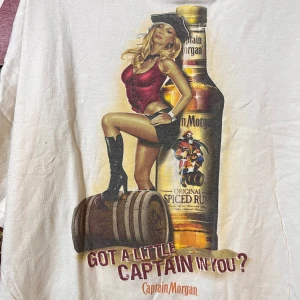 Vit vintage  t-shirt med Captain Morgan-tryck - Säljer en vit t-shirt med ett coolt Captain Morgan-tryck. Den har en bild av en flaska och en kvinna i röd topp och svarta stövlar. Perfekt för en avslappnad dag eller fest.