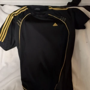 Svart tränings t-shirt från Adidas med gula detaljer - Svart tränings t-shirt från Adidas med ikoniska gula ränder på axlarna och gul logga på bröstet. Perfekt för sport och träning. Kortärmad design i ett bekvämt material.