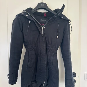 Svart parkajacka från Modström - Snygg svart parkajacka från Modström med dragkedja och knappar. Jackan har en huva och praktiska fickor framtill. Perfekt för kyligare dagar med stilrena detaljer som förstärkningar på armbågarna.