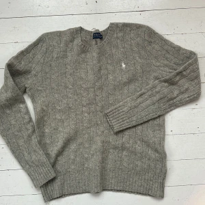 Grå kabelstickad tröja från Polo Ralph Lauren - Säljer en stilren grå kabelstickad tröja från Polo Ralph Lauren. Tröjan är tillverkad i en lyxig blandning av merinoull och kashmir, vilket gör den både mjuk och varm. Den har långa ärmar och en klassisk rund halsringning. Perfekt för kyliga dagar!