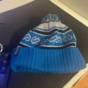 Blå och grå mössa med tofs - Snygg blå och grå mössa från Stellar med tofs på toppen. Den har ett mönster i blått och vitt samt en ribbad kant. Perfekt för kyliga dagar!