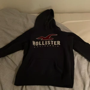 Svart hoodie från Hollister - Säljer en svart hoodie från Hollister med stor logga på framsidan. Tröjan har en klassisk känguruficka och en bekväm huva. Perfekt för en avslappnad stil.