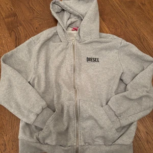 Grå hoodie från Diesel - Säljer en snygg grå hoodie från Diesel i storlek 16Y, som motsvarar xs. Den är i bomull och har en dragkedja framtill. Perfekt för höst och vår med sin bekväma passform och stilrena design. Diesel-loggan är tryckt på bröstet. Passar både till vardags och chill. Skick 8,5. Inga problem på tröjan. Priset kan diskuteras 😉