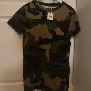 Säljer en supercool camouflage klänning från Moschino i storlek XS. Den är kortärmad och perfekt för en avslappnad stil. Materialet känns skönt mot huden och den har en klassisk rund hals. Perfekt för dig som vill sticka ut lite extra!