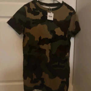 Camouflage klänning  från Moschino - Säljer en supercool camouflage klänning från Moschino i storlek XS. Den är kortärmad och perfekt för en avslappnad stil. Materialet känns skönt mot huden och den har en klassisk rund hals. Perfekt för dig som vill sticka ut lite extra!