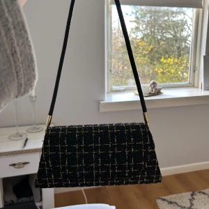 Svart rutig axelväska - Snygg svart axelväska med rutigt mönster och gulddetaljer. Perfekt för både vardag och fest! Väskan har en elegant design med en lång rem och stängs med magnetknapp. Den är i mycket bra skick och redo att användas.