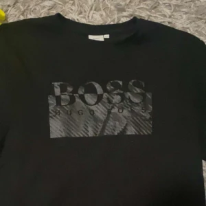 Svart t-shirt från Hugo Boss - Snygg svart t-shirt från Hugo Boss med stort BOSS-tryck på framsidan. Perfekt för en stilren och avslappnad look. Passar både till vardags och fest. Materialet känns mjukt och skönt mot huden.